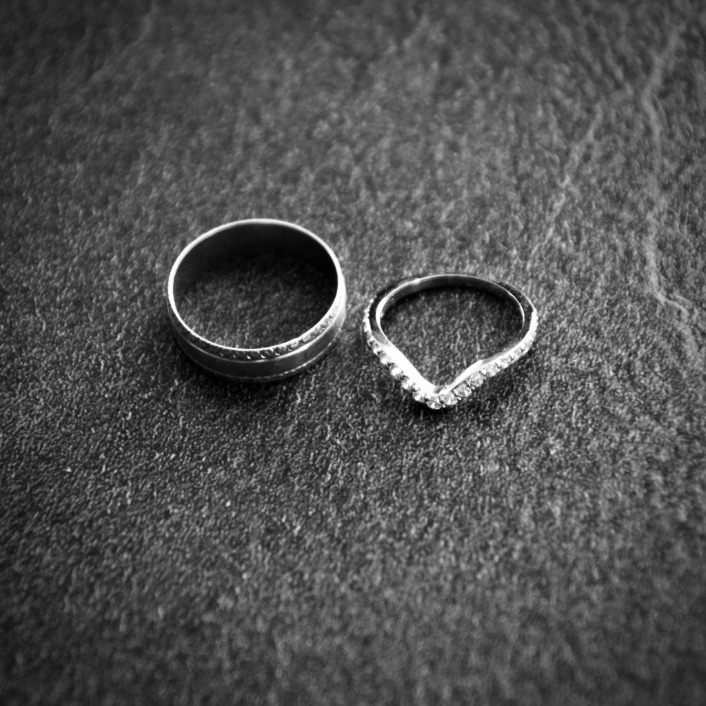 A close-up image of two silver rings on a textured surface. One is a plain band, and the other is a decorative ring with small stones arranged in a V shape.