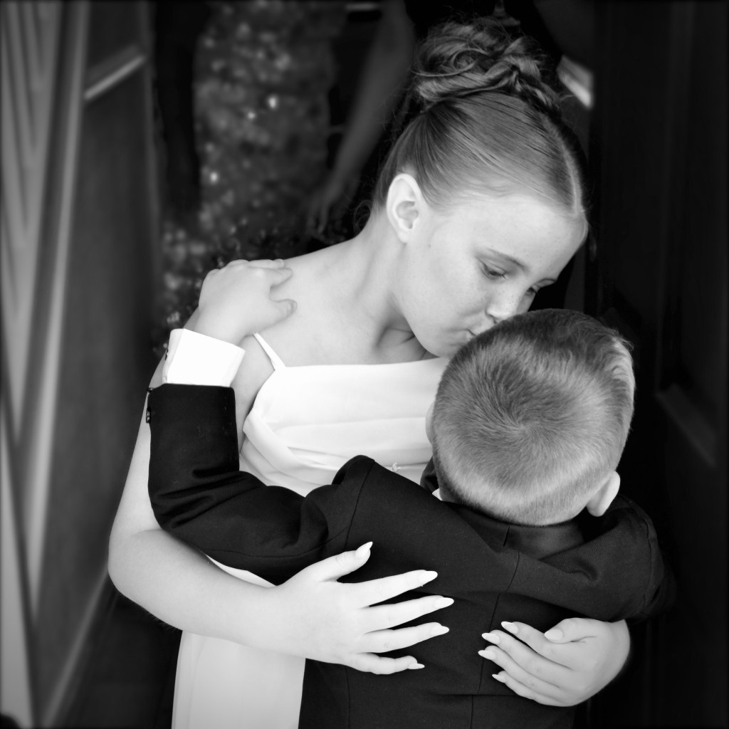 A girl in a white dress tenderly kisses a boy in formal attire, embracing him with affection. The image is in black and white, conveying a heartfelt moment.