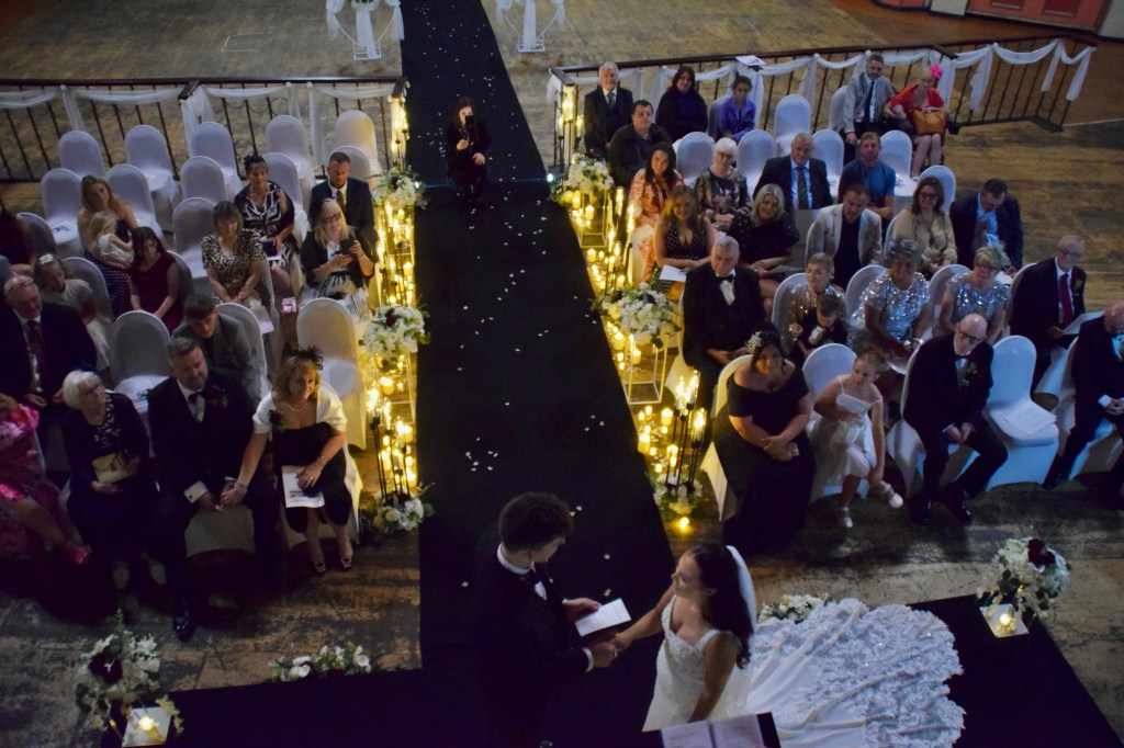 A wedding ceremony taking place indoors with guests seated on white chairs. The couple is standing on a black carpet surrounded by floral arrangements and candles. The atmosphere is elegant and celebratory.
