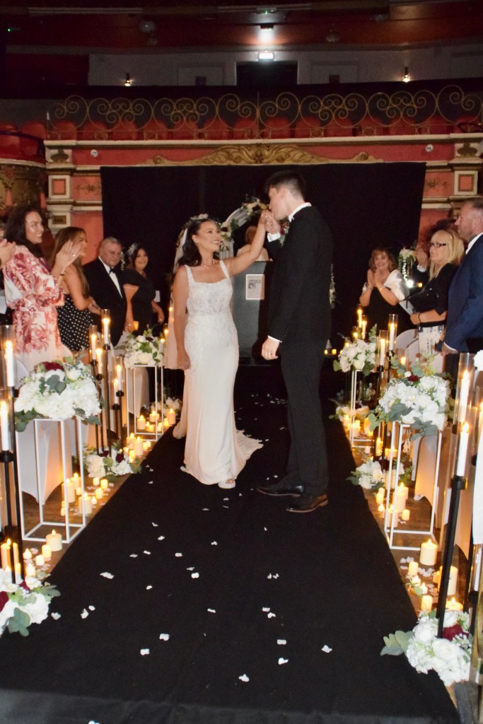 A bride and groom share a romantic moment during their wedding ceremony, surrounded by guests on either side, with a decorated aisle featuring flowers and candles.