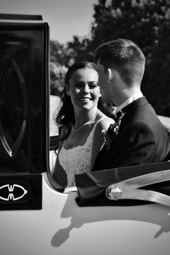 A newlywed couple shares a joyful moment inside a classic car, smiling at each other. The photo is in black and white, capturing the elegance of the wedding day.