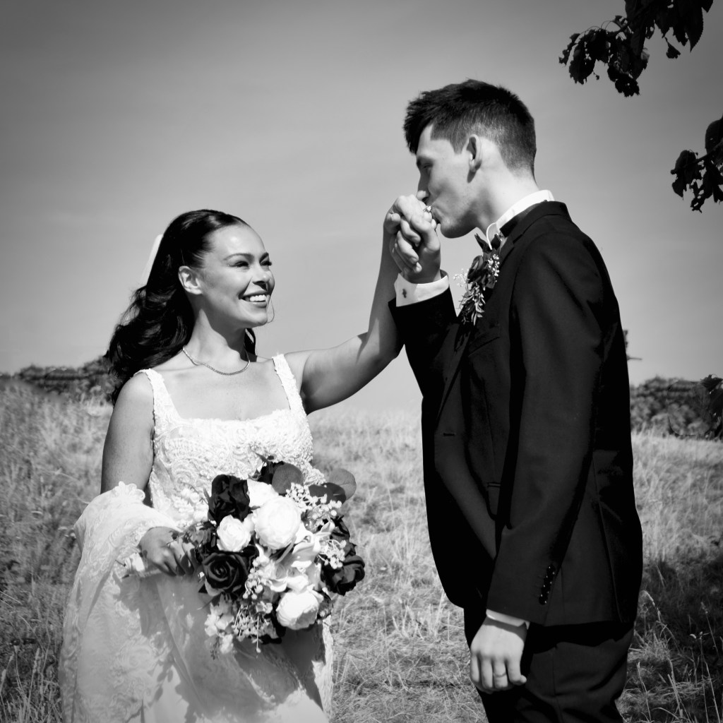 A happy bride in a white wedding dress holds a bouquet and smiles at her groom, who is kissing her hand in a romantic outdoor setting.