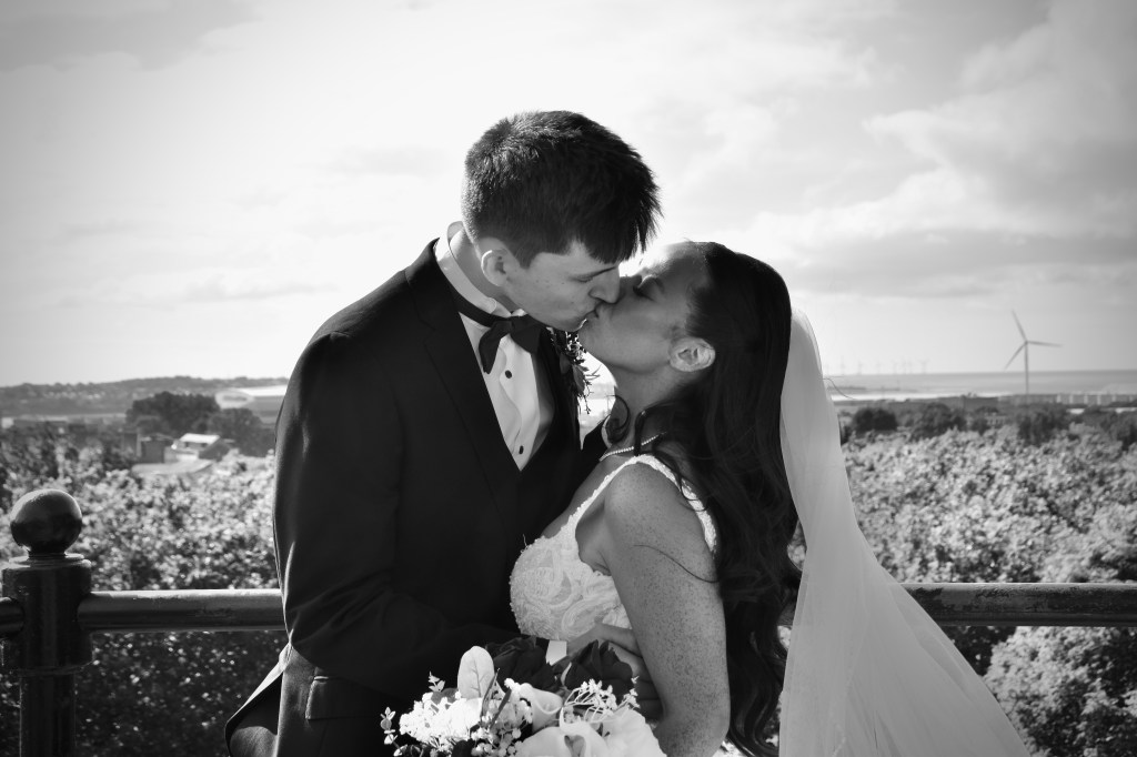 A newlywed couple sharing a romantic kiss outdoors, surrounded by a scenic landscape.