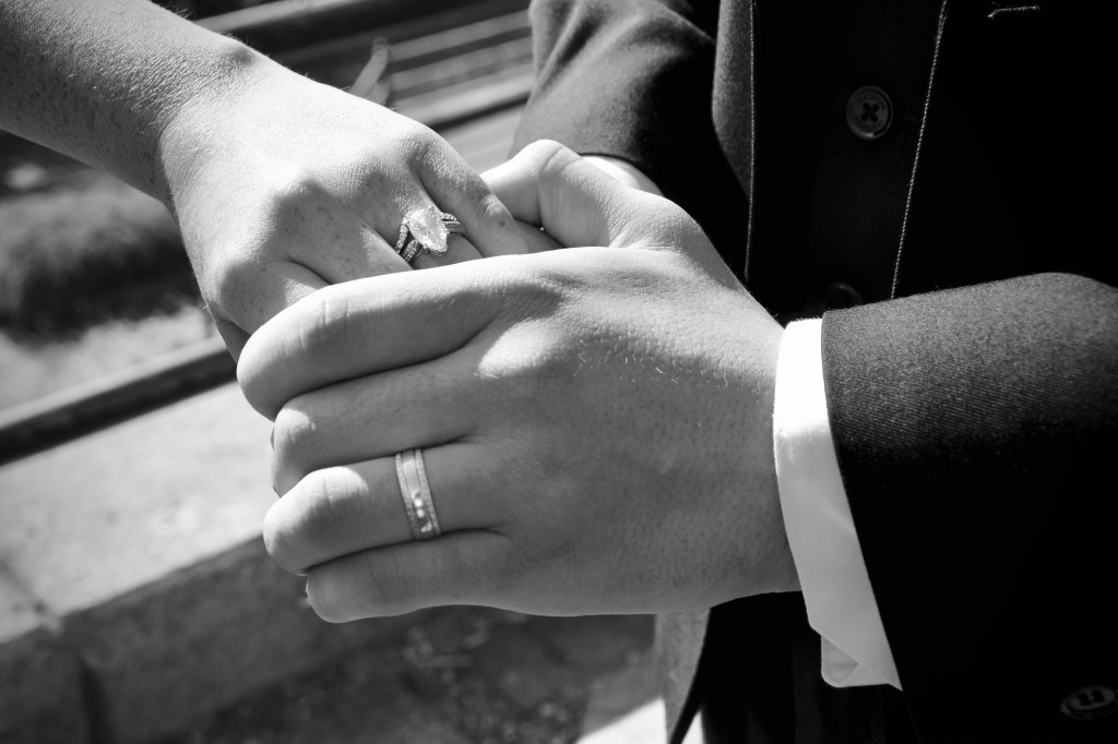 Close-up of two hands holding each other, one wearing a diamond engagement ring and the other a simple wedding band, against a blurred background.