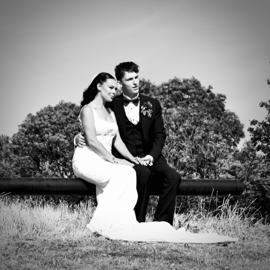 Black and white image of a bride and groom sitting closely together on a bench, surrounded by greenery.