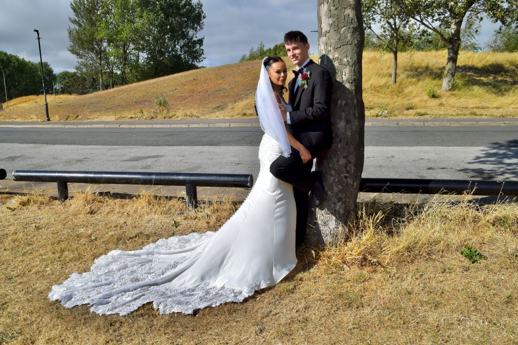 A bride and groom pose together outdoors, with the bride leaning against a tree and wearing a long white wedding gown. The groom stands beside her, dressed in a black suit, embracing her. The background features a grassy area and a road.