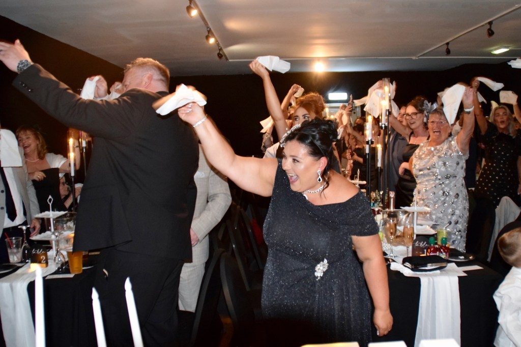 A lively celebration scene with a diverse group of people raising napkins in the air, showcasing joy and excitement at an event. A woman in a sparkly black dress is prominently smiling and participating in the festivities.