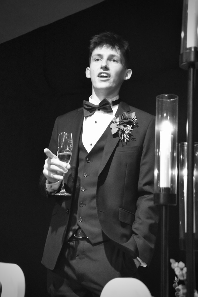 A young man in a tuxedo delivering a speech while holding a champagne glass