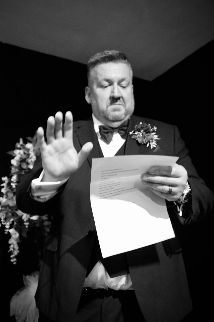 A man in formal attire holds a paper and gestures with his hand, appearing to give a speech or presentation.