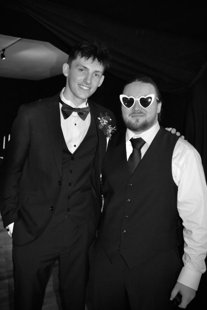 Two men posing together in formal attire, with one wearing heart-shaped sunglasses and the other in a tuxedo.