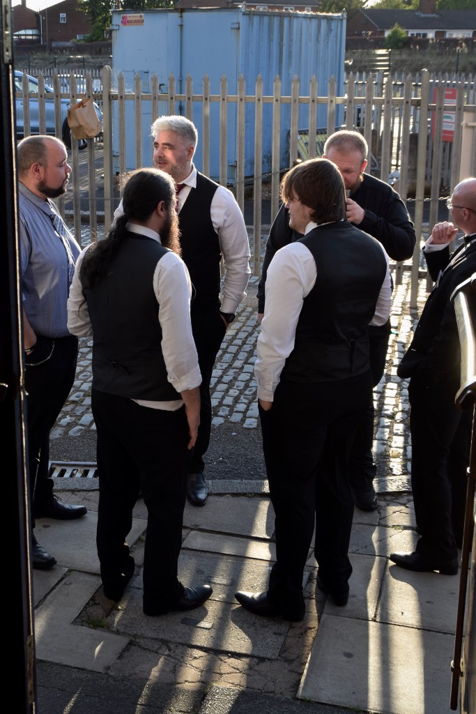A group of six men dressed in formal attire, gathered outside in the sunlight, engaging in conversation near a fence and a blue shipping container.