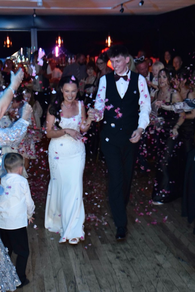 A bride and groom walking hand in hand through a crowd, surrounded by guests celebrating with rose petals in the air at a wedding reception.