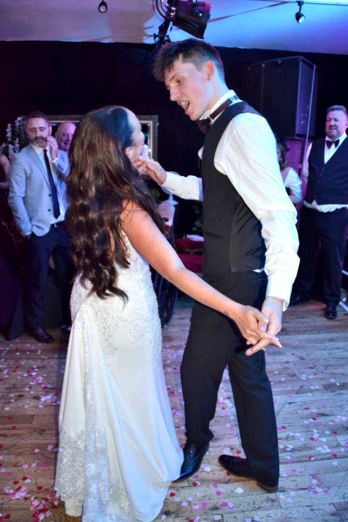 A bride and groom dancing together in a festive atmosphere, surrounded by wedding guests. The bride is wearing an elegant white dress with intricate detailing, and the groom is dressed in a formal black and white suit. The floor is scattered with pink and white rose petals.