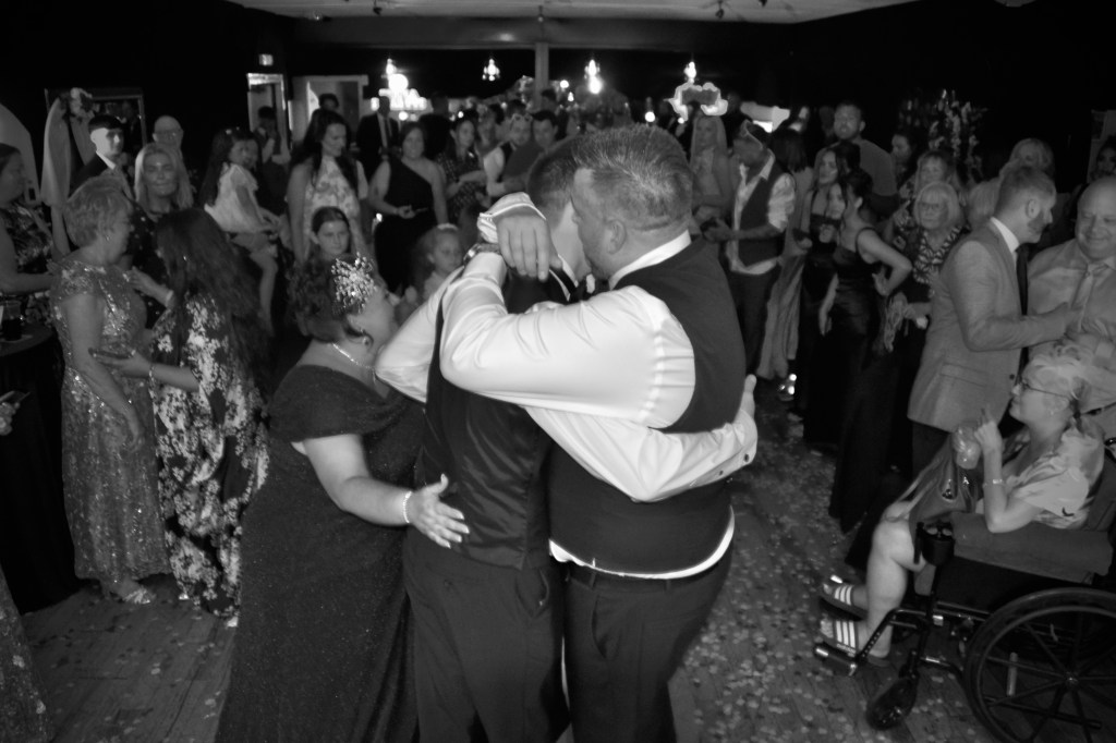 A joyful wedding reception scene in black and white, featuring a couple embracing while dancing, surrounded by guests enjoying the celebration, with decorations and a festive atmosphere.