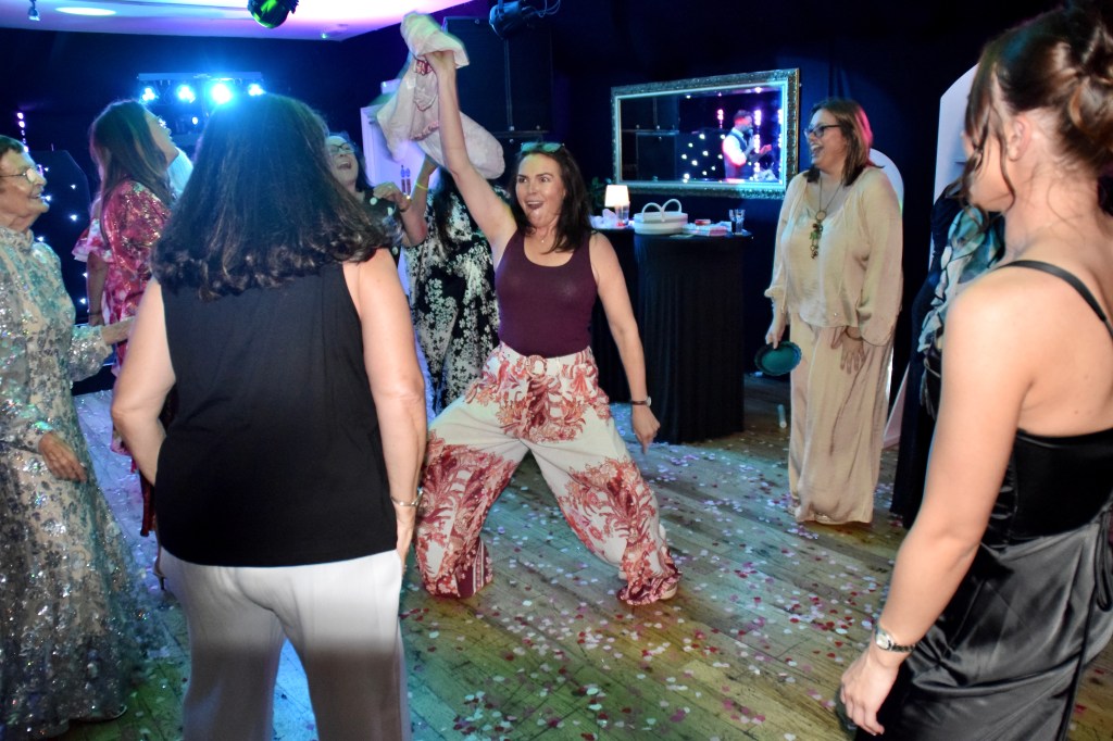 A lively indoor party scene with a group of women dancing. The woman in the foreground is striking a playful pose while holding a piece of clothing above her head. The background features colorful lights and confetti on the floor.